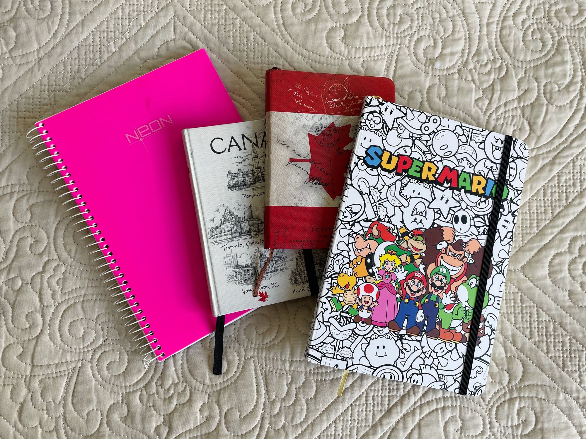 Um caderno rosa, seguido por dois pequenos cadernos com temática canadense e um caderno branco com capa do Super Mario World.